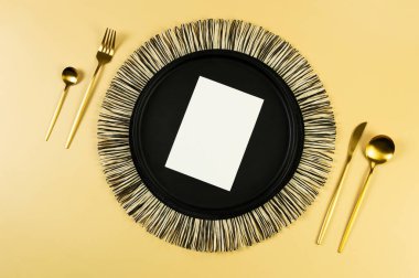 Siyah hasır masa paspası, siyah tabak, altın çatal bıçak takımı, sarı arka planda beyaz kart. Saman yuvarlak tepsi, düz konum, kopyalama alanı. 