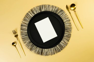 Siyah hasır masa paspası, siyah tabak, altın çatal bıçak takımı, sarı arka planda beyaz kart. Saman yuvarlak tepsi, düz konum, kopyalama alanı. 