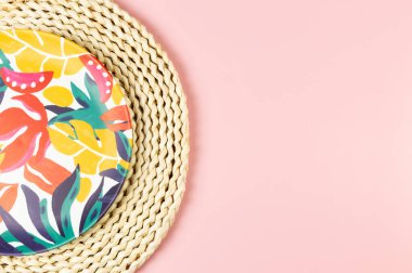 Kahverengi hasır masa paspasının üst görüntüsü, pembe arka planda renkli plaka. Saman tepsisi, egzotik bitki tabağı, yaz pikniği. Düz yat, boşluğu kopyala.