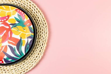 Kahverengi hasır masa paspası, siyah tepsi ve pembe arka planda renkli plaka. Saman tepsisi, egzotik bitki tabağı, yaz pikniği. Düz yat, boşluğu kopyala.