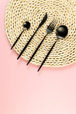 Kahverengi hasır masa paspası, pembe arka planda siyah çatal bıçak takımı. Saman yuvarlak tepsi, düz konum, kopyalama alanı.