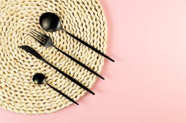 Kahverengi hasır masa paspası, pembe arka planda siyah çatal bıçak takımı. Saman yuvarlak tepsi, düz konum, kopyalama alanı.