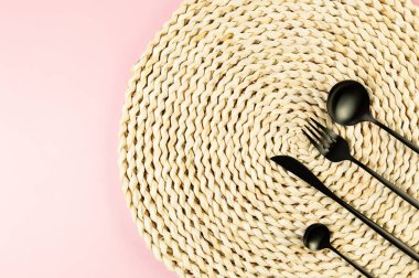 Kahverengi hasır masa paspası, pembe arka planda siyah çatal bıçak takımı. Saman yuvarlak tepsi, düz konum, kopyalama alanı.