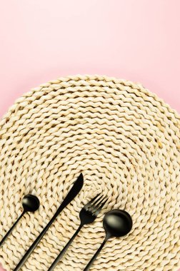 Kahverengi hasır masa paspası, pembe arka planda siyah çatal bıçak takımı. Saman yuvarlak tepsi, düz konum, kopyalama alanı.