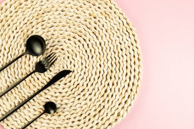 Kahverengi hasır masa paspası, pembe arka planda siyah çatal bıçak takımı. Saman yuvarlak tepsi, düz konum, kopyalama alanı.