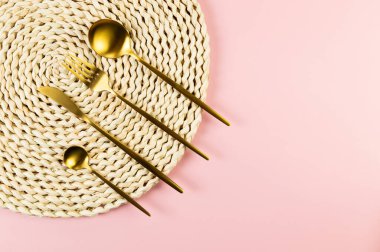 Kahverengi hasır masa paspası, pembe arka planda altın çatal bıçak seti. Saman yuvarlak tepsi, düz konum, kopyalama alanı.