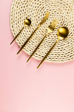 Kahverengi hasır masa paspası, pembe arka planda altın çatal bıçak seti. Saman yuvarlak tepsi, düz konum, kopyalama alanı.