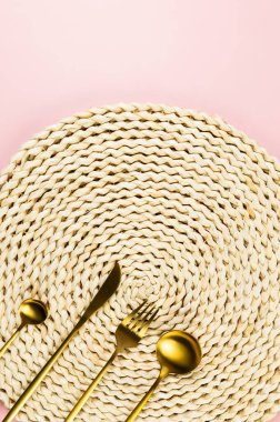 Kahverengi hasır masa paspası, pembe arka planda altın çatal bıçak seti. Saman yuvarlak tepsi, düz konum, kopyalama alanı.