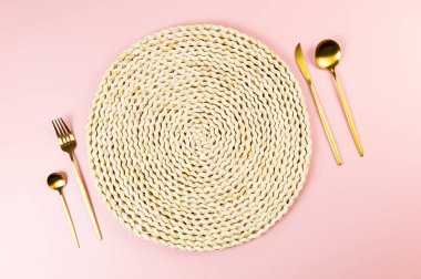 Kahverengi hasır masa paspası, pembe arka planda altın çatal bıçak seti. Saman yuvarlak tepsi, düz konum, kopyalama alanı.