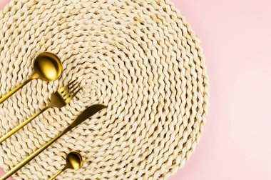 Kahverengi hasır masa paspası, pembe arka planda altın çatal bıçak seti. Saman yuvarlak tepsi, düz konum, kopyalama alanı.