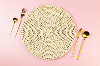 Kahverengi hasır masa paspası, pembe arka planda altın çatal bıçak seti. Saman yuvarlak tepsi, düz konum, kopyalama alanı.