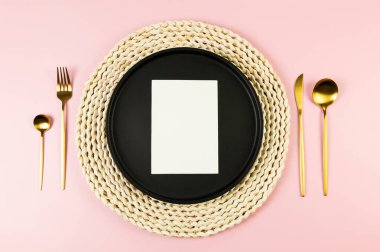 Kahverengi hasır masa paspası, siyah tabak, pembe arka planda altın çatal bıçak. Beyaz kart, saman yuvarlak tepsi, düz konum, kopyalama alanı.