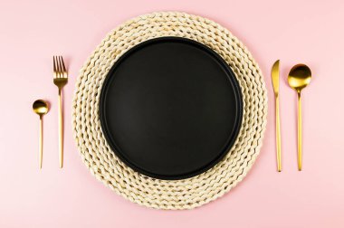 Kahverengi hasır masa paspası, siyah tabak, pembe arka planda altın çatal bıçak. Saman yuvarlak tepsi, düz konum, kopyalama alanı.