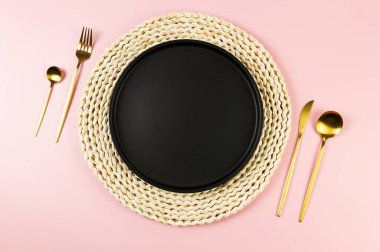 Kahverengi hasır masa paspası, siyah tabak, pembe arka planda altın çatal bıçak. Saman yuvarlak tepsi, düz konum, kopyalama alanı.
