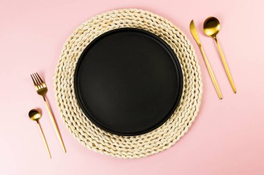 Kahverengi hasır masa paspası, siyah tabak, pembe arka planda altın çatal bıçak. Saman yuvarlak tepsi, düz konum, kopyalama alanı.
