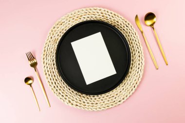 Kahverengi hasır masa paspası, siyah tabak, pembe arka planda altın çatal bıçak. Beyaz kart, saman yuvarlak tepsi, düz konum, kopyalama alanı.