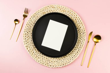 Kahverengi hasır masa paspası, siyah tabak, pembe arka planda altın çatal bıçak. Beyaz kart, saman yuvarlak tepsi, düz konum, kopyalama alanı.