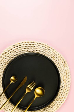 Kahverengi hasır masa paspası, siyah tabak, pembe arka planda altın çatal bıçak. Saman yuvarlak tepsi, düz konum, kopyalama alanı.