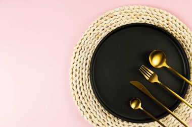 Kahverengi hasır masa paspası, siyah tabak, pembe arka planda altın çatal bıçak. Saman yuvarlak tepsi, düz konum, kopyalama alanı.