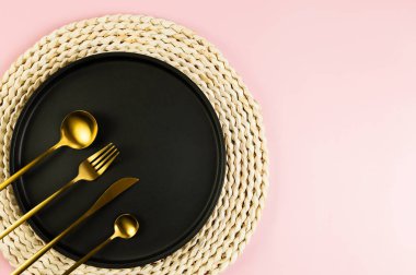Kahverengi hasır masa paspası, siyah tabak, pembe arka planda altın çatal bıçak. Saman yuvarlak tepsi, düz konum, kopyalama alanı.