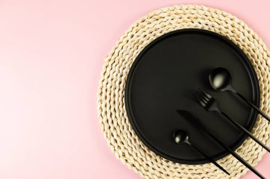 Kahverengi hasır masa paspası, siyah tabak, pembe arka planda siyah çatal bıçak. Saman yuvarlak tepsi, düz konum, kopyalama alanı.