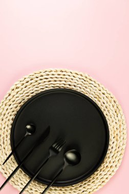 Kahverengi hasır masa paspası, siyah tabak, pembe arka planda siyah çatal bıçak. Saman yuvarlak tepsi, düz konum, kopyalama alanı.