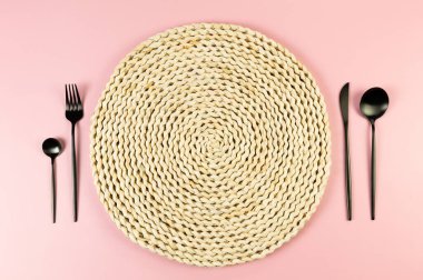 Kahverengi hasır masa paspası, pembe arka planda siyah çatal bıçak takımı. Saman yuvarlak tepsi, düz konum, kopyalama alanı.