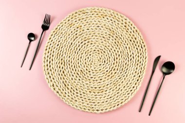 Kahverengi hasır masa paspası, pembe arka planda siyah çatal bıçak takımı. Saman yuvarlak tepsi, düz konum, kopyalama alanı.