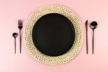 Kahverengi hasır masa paspası, siyah tabak, pembe arka planda siyah çatal bıçak. Saman yuvarlak tepsi, düz konum, kopyalama alanı.