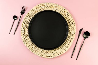 Kahverengi hasır masa paspası, siyah tabak, pembe arka planda siyah çatal bıçak. Saman yuvarlak tepsi, düz konum, kopyalama alanı.