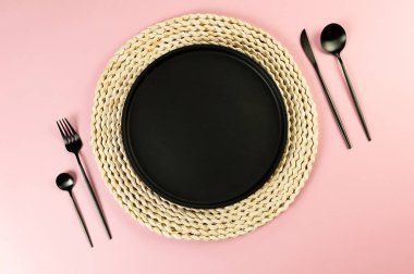 Kahverengi hasır masa paspası, siyah tabak, pembe arka planda siyah çatal bıçak. Saman yuvarlak tepsi, düz konum, kopyalama alanı.