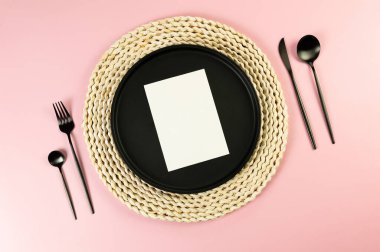 Kahverengi hasır masa paspası, siyah tabak, pembe arka planda siyah çatal bıçak. Beyaz kart, saman yuvarlak tepsi, düz konum, kopyalama alanı.