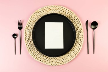 Kahverengi hasır masa paspası, siyah tabak, pembe arka planda siyah çatal bıçak. Beyaz kart, saman yuvarlak tepsi, düz konum, kopyalama alanı.