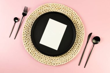 Kahverengi hasır masa paspası, siyah tabak, pembe arka planda siyah çatal bıçak. Beyaz kart, saman yuvarlak tepsi, düz konum, kopyalama alanı.