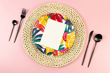 Kahverengi hasır masa paspası, renkli tabak, pembe arka planda siyah çatal bıçak. Beyaz kart, saman yuvarlak tepsi, egzotik bitki tabağı, yaz pikniği. Düz yat, boşluğu kopyala.