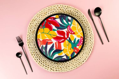 Kahverengi hasır masa paspası, renkli tabak, pembe arka planda siyah çatal bıçak. Siyah tepsi, saman yuvarlak tepsi, egzotik bitki tabağı, yaz pikniği. Düz yat, boşluğu kopyala.