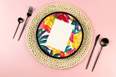 Kahverengi hasır masa paspası, renkli tabak, pembe arka planda siyah çatal bıçak. Siyah tepsi, beyaz kart, saman yuvarlak tepsi, egzotik bitki plakası. Yaz pikniği, düz yatak, kopyalama alanı.