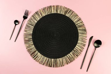 Siyah hasır masa paspası, pembe arka planda siyah çatal bıçak takımı. Saman yuvarlak tepsi, düz konum, kopyalama alanı.