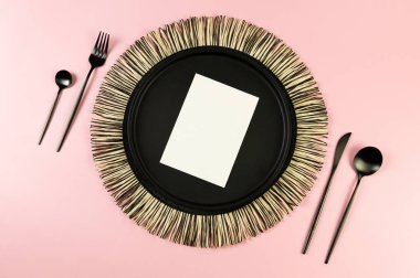 Siyah hasır masa paspası, siyah çatal bıçak takımı, pembe arka planda beyaz kart. Saman yuvarlak tepsi, düz konum, kopyalama alanı.