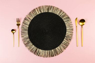 Siyah hasır masa paspası, pembe arka planda altın çatal bıçak takımı. Saman yuvarlak tepsi, düz konum, kopyalama alanı.