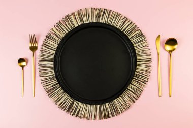 Siyah hasır masa paspası, siyah tabak, pembe arka planda altın çatal bıçak. Saman yuvarlak tepsi, düz konum, kopyalama alanı.