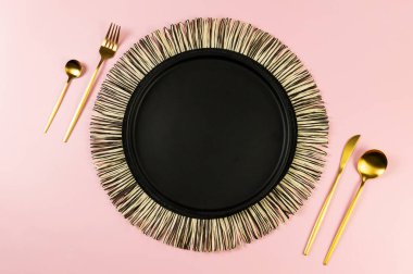 Siyah hasır masa paspası, siyah tabak, pembe arka planda altın çatal bıçak. Saman yuvarlak tepsi, düz konum, kopyalama alanı.