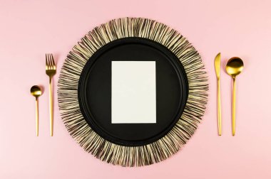 Siyah hasır masa paspası, siyah tabak, altın çatal bıçak takımı, pembe arka planda beyaz kart. Saman yuvarlak tepsi, düz konum, kopyalama alanı.