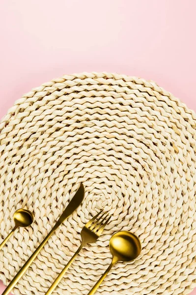 Kahverengi hasır masa paspası, pembe arka planda altın çatal bıçak seti. Saman yuvarlak tepsi, düz konum, kopyalama alanı.