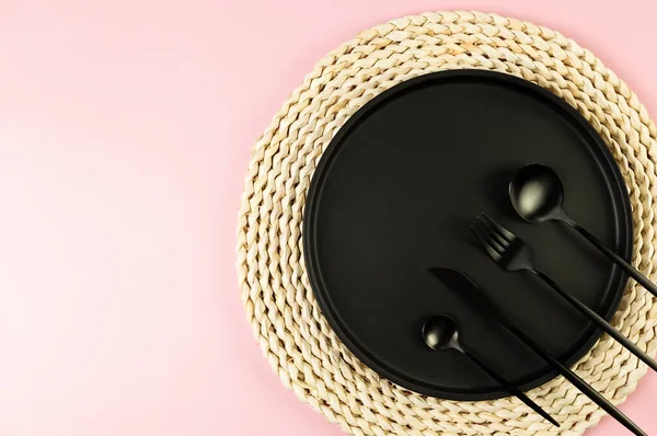 Kahverengi hasır masa paspası, siyah tabak, pembe arka planda siyah çatal bıçak. Saman yuvarlak tepsi, düz konum, kopyalama alanı.