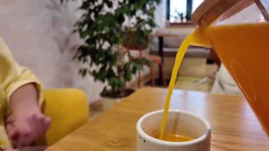 sarı süveter eli portakal suyunu benekli seramik bardağa döküyor rahat kafeteryadaki ahşap masaya, saksı ve yumuşak sandalyeli, doğal ışık, dingin