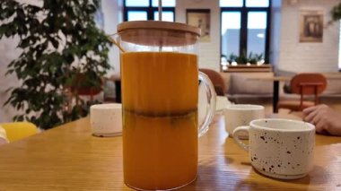 Ahşap masada cam sürahi çayı, elle karıştırılmış kaşık, cam kenarında benekli seramik bardak, saksı bitkisi ve büyük pencereli güneş ışığı kahvesi, sıcak kehribar.