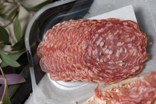 Stacked sliced salami in packaging, vacuumpacked transparent container showing thin marbled cold cuts on parchment, retail shelf aesthetic with label