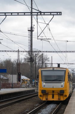 Sarı tren, bulutlu gökyüzünün altındaki platforma yaklaşıyor, üstten kablolar ve sinyal direkleri sahne, platform kenarı ve ıslak raylar ışığı yansıtıyor, uzak şehir