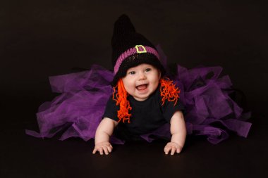 Cadılar Bayramı cadı kostümü giyen mutlu bir bebek. Turuncu iplik saçlı, siyah şapkalı ve mor tüllü etekli. Stüdyoda siyah arka planda gülüp sürünüyordu. Cadılar Bayramı ve sonbahar temalı içerik için şenlikli ve eğlenceli görüntü.
